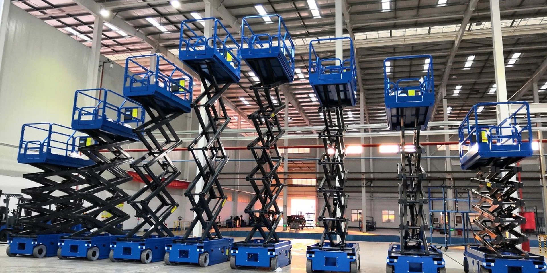Ocho plataformas de tijera azules con la plataforma elevada, alineadas en una nave industrial