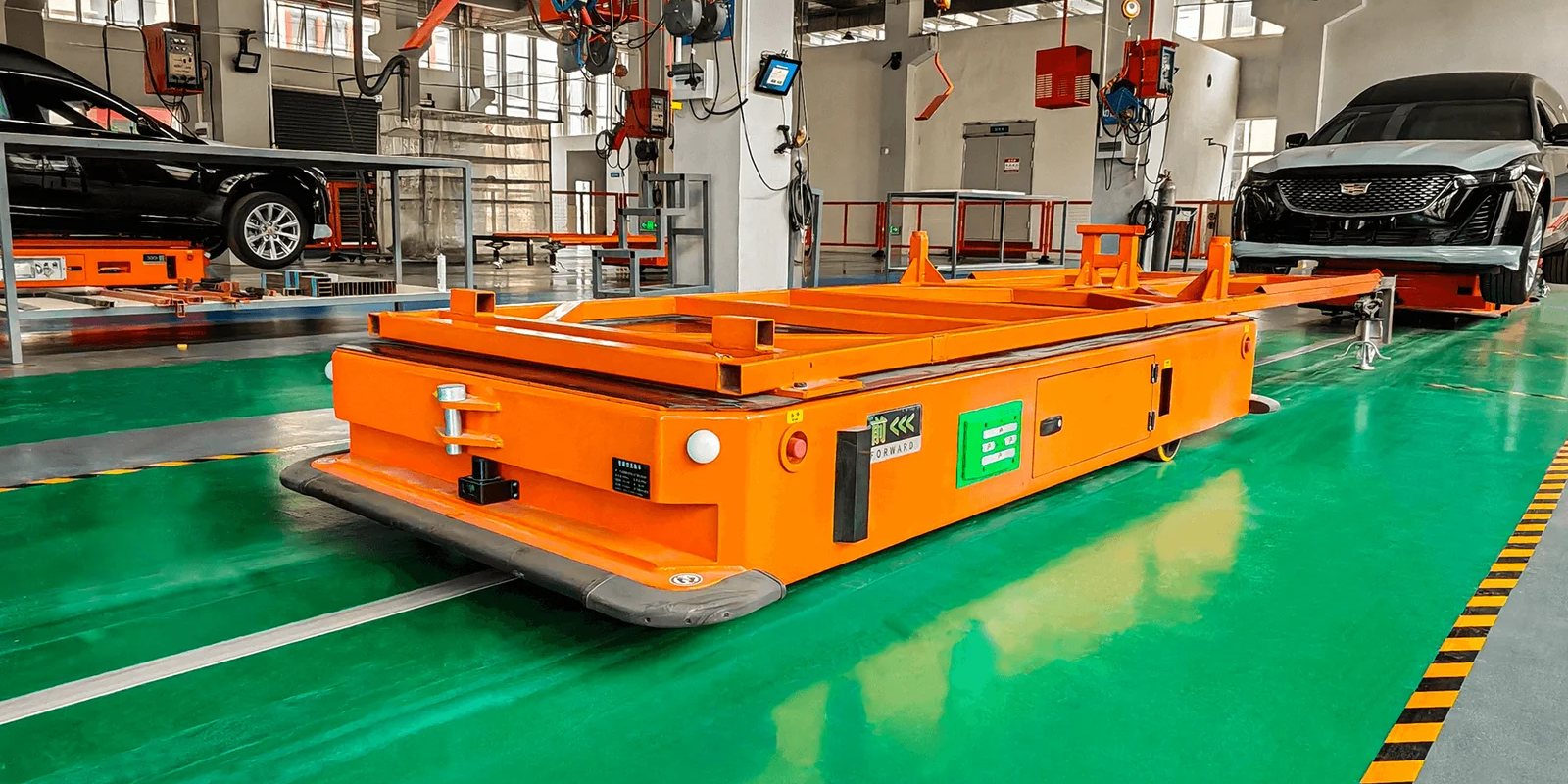 Orange automated guided vehicle on a green factory floor, with car bodies on the line behind it
